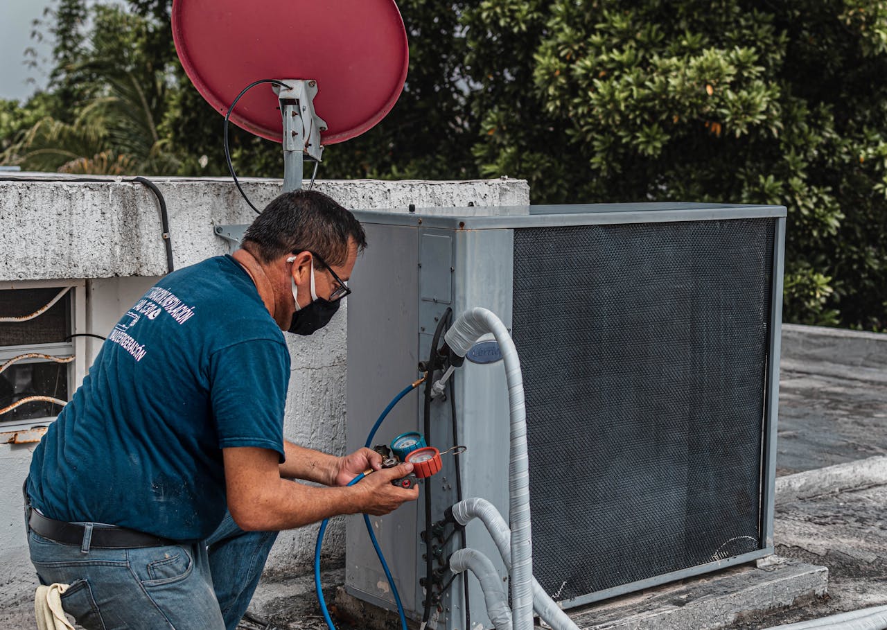 gallery-1 Technician repairing an air conditioner unit outdoors, wearing a facemask and using a manifold gauge.